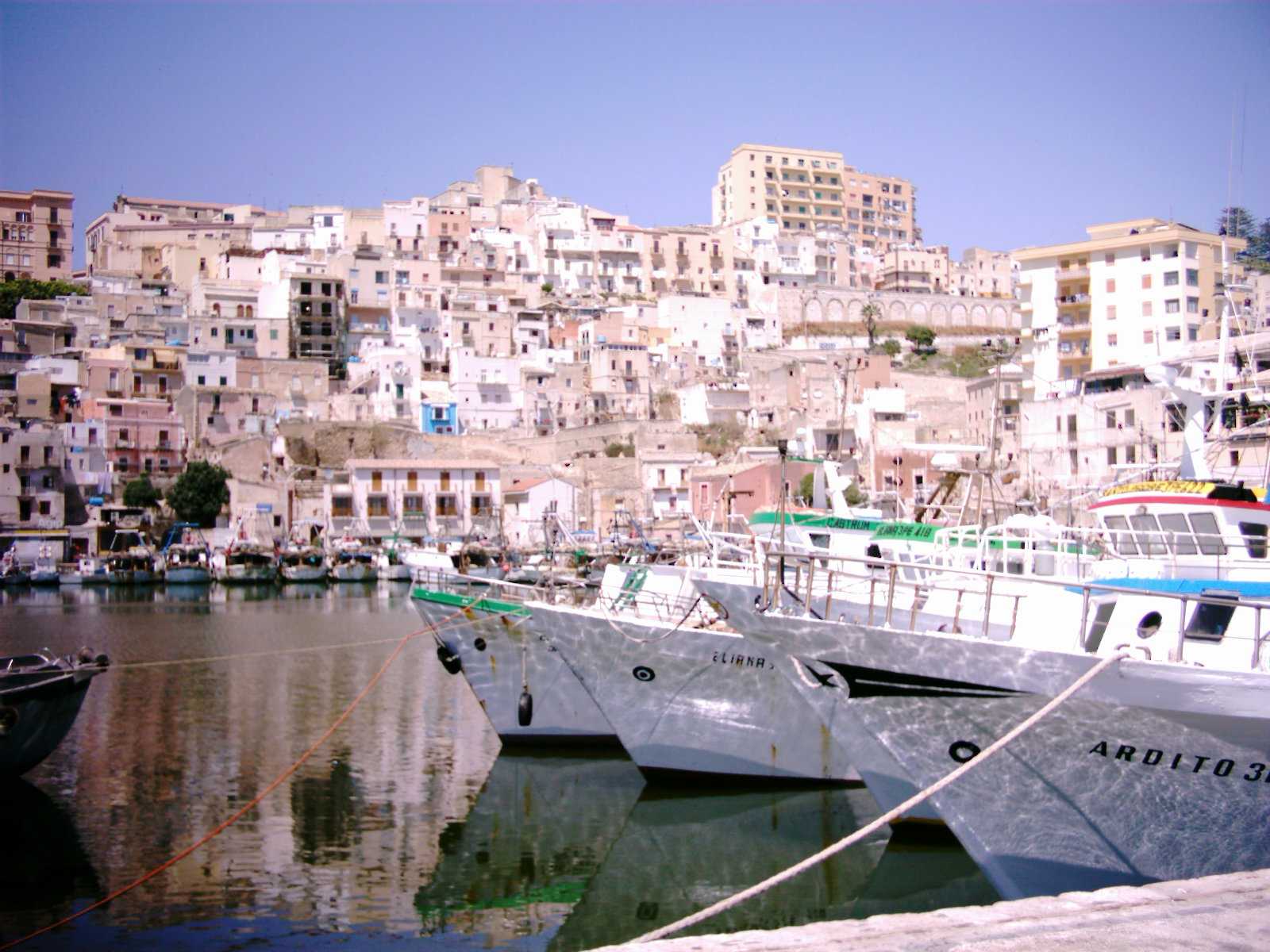 The harbor at Sciacca, Sicily, with buildings rising from the waterfront and fishing boats moored below.